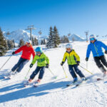 Famille (deux adultes, deux enfants) skie joyeusement sur une piste ensoleillée. Montagnes enneigées et télésièges à l'arrière-plan.