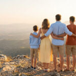 Famille de 4 (parents, 2 enfants) dos à la caméra sur un sommet rocheux, admirant un vaste paysage montagneux au soleil couchant.