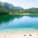 Vue aérienne du lac d'Aiguebelette avec ses eaux émeraude et bleues, entouré de montagnes boisées, et une plage de sable avec des baigneurs.