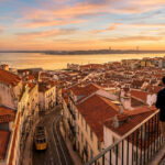 Vue aérienne de Lisbonne au coucher du soleil. Des personnes contemplent la ville, le Tage, le Pont du 25 Avril et un tram jaune.