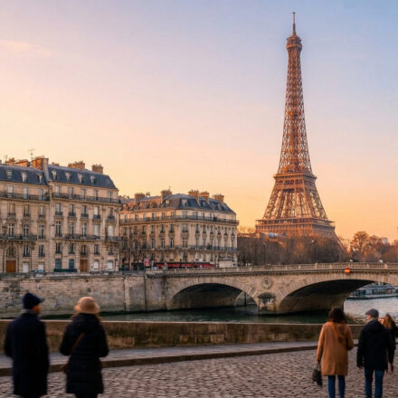 La Tour Eiffel illuminée au coucher du soleil, un pont sur la Seine, des bâtiments haussmanniens et des passants sur un quai pavé.