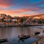 Vue panoramique de Porto au coucher du soleil, avec le pont Dom Luís I enjambant le Douro, des rabelos et les façades colorées de la Ribeira.