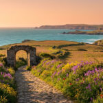 Chemin de pierre bordé de fleurs violettes et jaunes menant à une arche de pierre, mer turquoise et falaises en arrière-plan.