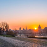 Lever de soleil vibrant sur Paris, montrant la Seine embrumée, les quais bordés d'arbres et les toits parisiens sous un ciel orange et bleu.