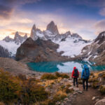 Comment visiter El Chaltén en 2026 : le guide complet Deux randonneurs marchent sur un sentier rocheux vers le mont Fitz Roy enneigé et le lac Sucia turquoise, sous un ciel orangé.