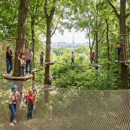 Accrobranche à Sannois : guide pour votre aventure nature Plusieurs familles font de l'accrobranche entre des arbres verdoyants, avec des ponts de cordes et filets. La Tour Eiffel est visible en arrière-plan.