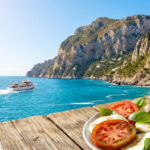 Trouver le meilleur parc d’attraction pour vos vacances Salade Caprese sur table en bois, surplombant la mer Méditerranée et les falaises de Capri avec un bateau naviguant.