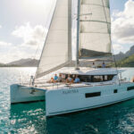 Catamaran blanc "AURORA" avec voiles hissées, naviguant sur mer turquoise. Passagers détendus. Montagnes tropicales en arrière-plan.