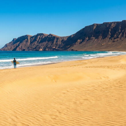 Un surfeur en combinaison marche sur une plage de sable doré avec des dunes, face à l'océan turquoise et des montagnes volcaniques.