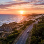 Coucher de soleil flamboyant sur la côte atlantique. L'océan, une plage, des falaises, une route sinueuse avec une voiture et des maisons parmi les pins.
