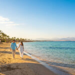 Hurghada est-il dangereux ? Sécurité et conseils pour 2026 Couple marchant main dans la main sur une plage de sable doré à Hurghada, bordée de palmiers, avec une mer turquoise et des montagnes.