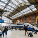 Intérieur de la Gare de Lyon avec un TGV à quai, des voyageurs, et le restaurant historique Le Train Bleu sous une verrière.