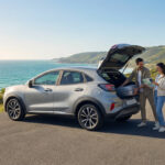 Couple souriant chargeant un SUV gris sur une route côtière ensoleillée avec l'océan turquoise et des collines verdoyantes en arrière-plan.