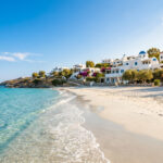 Où partir en juin : les meilleures destinations au soleil Vue d'une plage de sable blanc aux eaux turquoise, bordée de maisons blanches traditionnelles avec des toits bleus en Grèce.