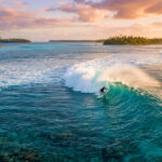Découvrir Corralejo aux Canaries : dunes, surf et volcans Surfeur sur une vague turquoise au cœur d'un lagon tropical, avec des îles bordées de palmiers sous un ciel pastel.