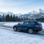 SUV bleu avec chaînes sur route sinueuse enneigée, montagnes et forêts majestueuses en arrière-plan sous un ciel clair.